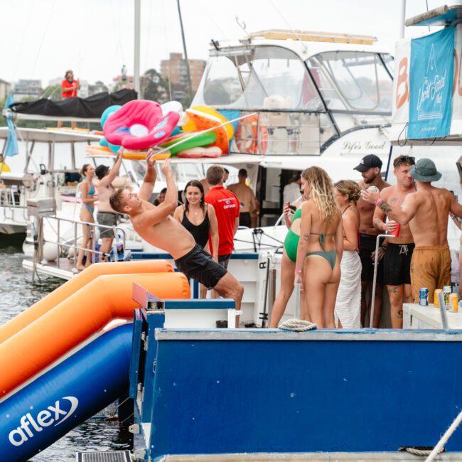 A lively group of people in swimsuits gathered on a docked boat, partying and socializing. A man holds up an inflatable float, while others enjoy drinks. Various boats and inflatables are visible in the background, indicating a fun aquatic setting with The Yacht Social Club Sydney Boat Hire.
