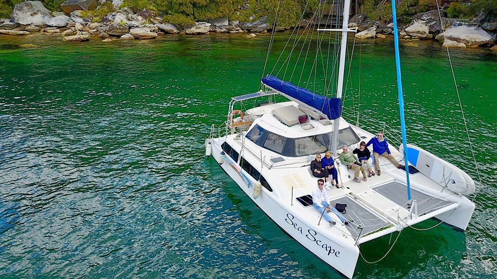 A group of people enjoying their time on the catamaran sailboat "Sara Scape" in a serene, green body of water, framed by rocky shorelines and trees. The boat, displaying elegant blue and white features along with its multiple sails, adds to the captivating scenery.