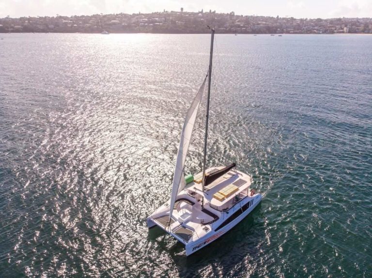 Aerial view of a sailboat gliding on a calm, glistening sea with a distant coastline and scattered buildings visible under a hazy sky. The boat's sails are partly unfurled, revealing the spacious deck.
