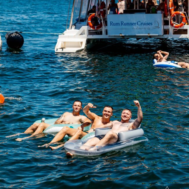 Three men float on inflatable loungers in a sunny water body, facing the camera with arms raised in a festive pose. Behind them is a white boat with a "Rum Runner Cruises" banner. Other people enjoy the water near the boat, creating an idyllic scene at The Yacht Social Club Sydney Boat Hire event.