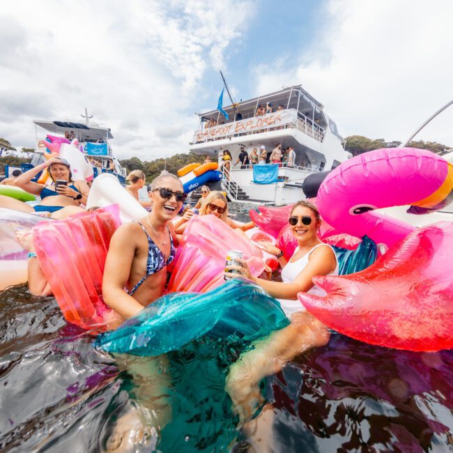 A group of people enjoys a sunny day on the water with large inflatable floats shaped like flamingos and mermaids. They smile and laugh near a boat named "Serenity Explorer," set against blue skies and trees, showcasing Boat Parties Sydney The Yacht Social Club.