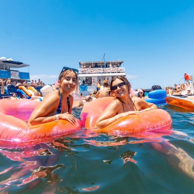 Two people are smiling and lounging on pink inflatable floats in the water, surrounded by numerous other individuals enjoying a sunny day. Several yachts and a floating platform with a slide are in the background, while the clear blue sky enhances the vibrant scene.