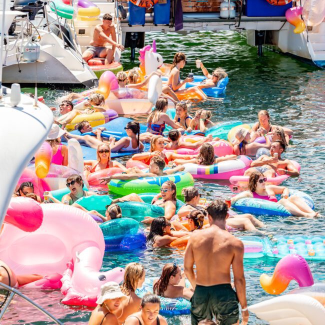 A large group of people enjoying a sunny day on the water, floating on colorful inflatables like unicorns and pink flamingos. Luxury yachts from The Yacht Social Club are docked nearby, and people are socializing, swimming, and lounging in swimsuits. The atmosphere is lively and festive.