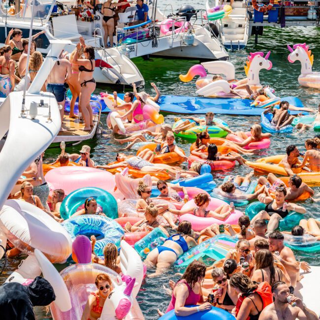 A lively scene at a lake party with numerous people on inflatable pool floats, including unicorns and donuts, gathered around several boats. Attendees are socializing, sunbathing, and enjoying the water in colorful attire under a bright, sunny sky—a perfect setting for The Yacht Social Club Event Boat Charters.