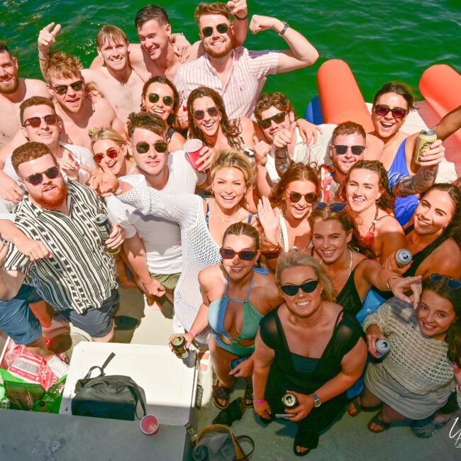 A large group of people in casual summer attire are smiling and posing for a photo on a boat with a green water background. Some are holding drinks and making playful gestures, while inflatable toys float nearby. A cooler with supplies is visible, enhancing the festive and jovial mood.