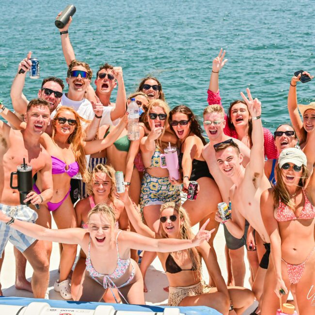 A group of about sixteen people, wearing swimsuits, are joyfully posing on a boat on a sunny day. They are smiling, raising their arms, and holding drinks, capturing the fun and excitement of a party or celebration on the water.