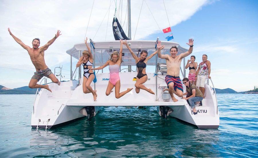 A group of six people joyfully jumping off a white catamaran into the azure sea. Everyone is wearing colorful swimsuits, and the boat proudly sports a flag. The scene unfolds on a sunny day with clear skies and calm waters, perfect for an unforgettable adventure.