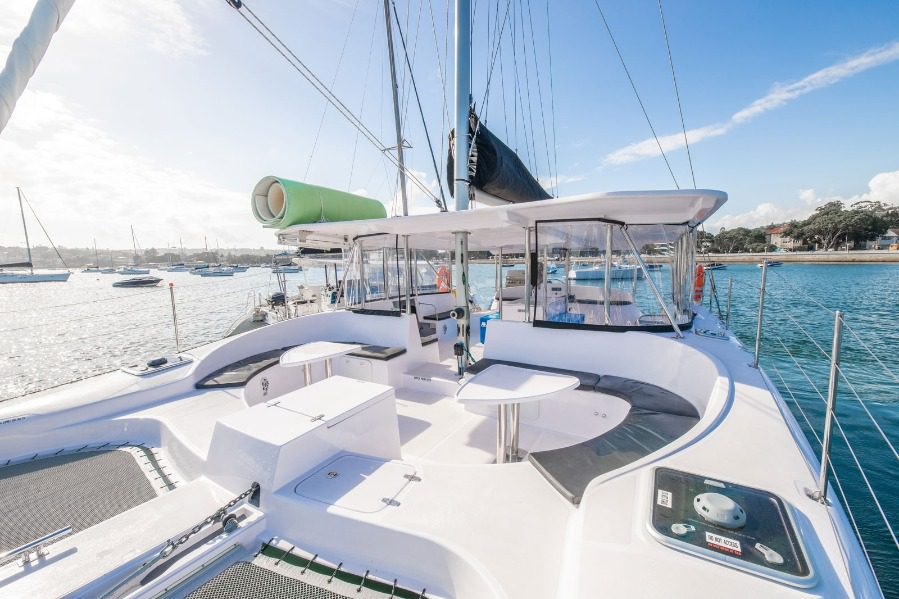 Caption: Spacious deck of a white catamaran yacht with seating areas and tables, anchored in a calm blue marina. Visible in the background are other boats and a shoreline with lush greenery under a clear, sunny sky. The inviting ambiance makes it perfect for relaxation or entertaining guests.