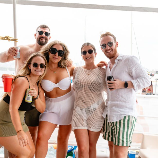 A group of five people poses on a boat deck from The Yacht Social Club Sydney Boat Hire. Four of them are wearing sunglasses and beachwear, while one person at the back is shirtless. They are smiling and holding drinks. The sky and water are visible in the background.