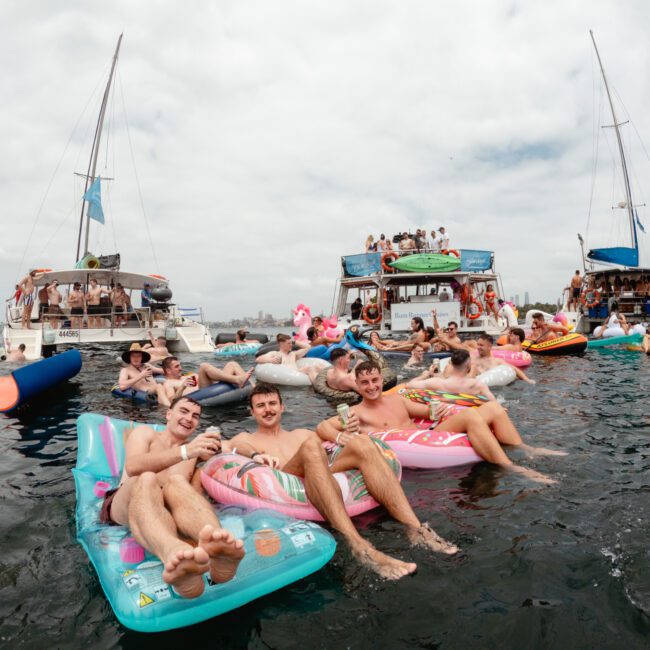 A group of people relaxing on colorful inflatable floaties in the water, surrounded by boats. They are enjoying a sunny day with drinks, while others are on the boats in the background. The atmosphere is festive and lively, embodying the spirit of The Yacht Social Club Sydney Boat Hire experience.