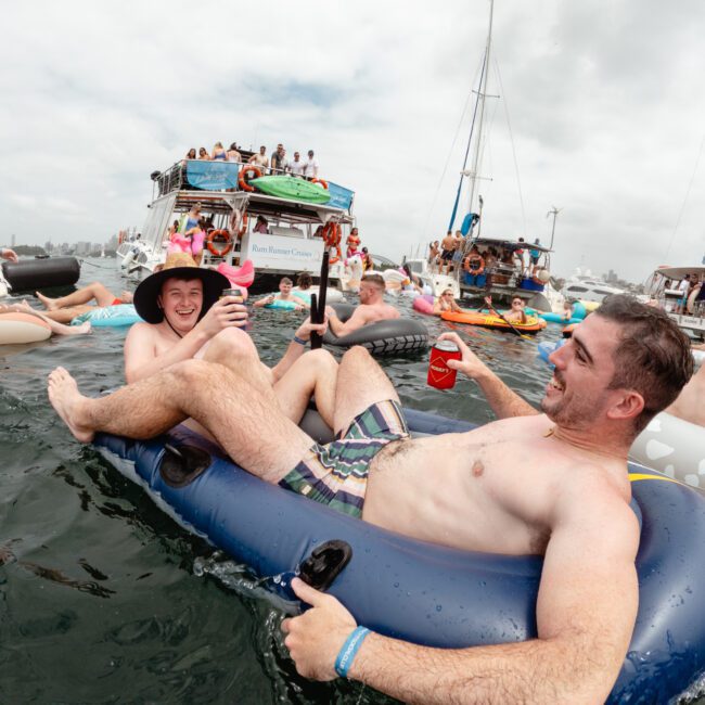 Two men relax in an inflatable pool float, smiling and holding drinks, surrounded by people on similar inflatables in the water. Boats and a large party on a luxury yacht are visible in the background under an overcast sky. The atmosphere is festive and cheerful, reminiscent of Boat Parties Sydney The Yacht Social Club.