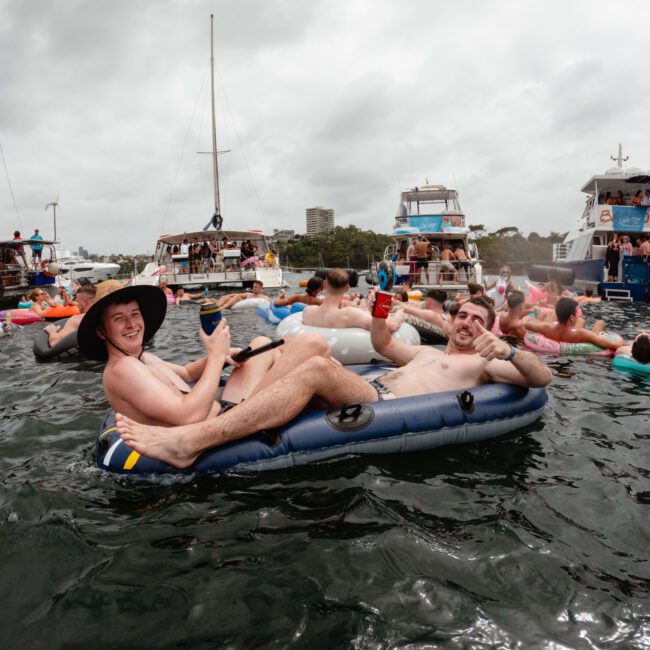 Two men are lounging on an inflatable raft in a crowded water area, holding drinks and smiling at the camera. Surrounding them are various people on rafts and in the water. In the background, several boats from The Yacht Social Club are anchored, and the sky is overcast.