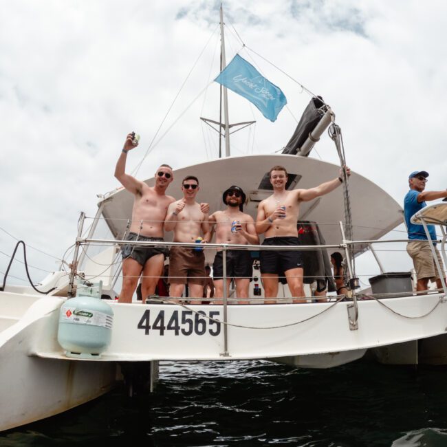 Four men, standing shirtless on the deck of a boat, smiling and holding drinks. A blue flag flies above them. Another person is visible on the right side of the image, standing at the helm. The boat's registration number "444565" is visible on the hull during a Yacht Social Club Sydney Boat Hire event.