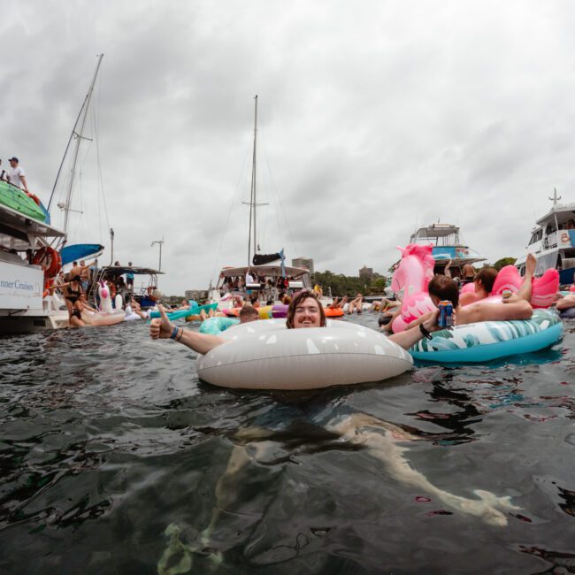 A man floating on an inflatable tube in the water, surrounded by others on various inflatables and boats. The sky is overcast, with several boats anchored in the background as people enjoy a gathering. It looks like a perfect day for Boat Parties Sydney The Yacht Social Club.