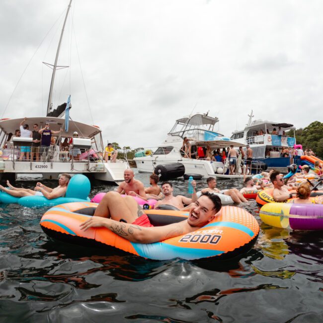 People gather and relax on colorful inflatable floats in the water near several anchored boats. A festive atmosphere is evident as individuals enjoy The Yacht Social Club event. The sky is overcast, adding a muted backdrop to the lively scene.