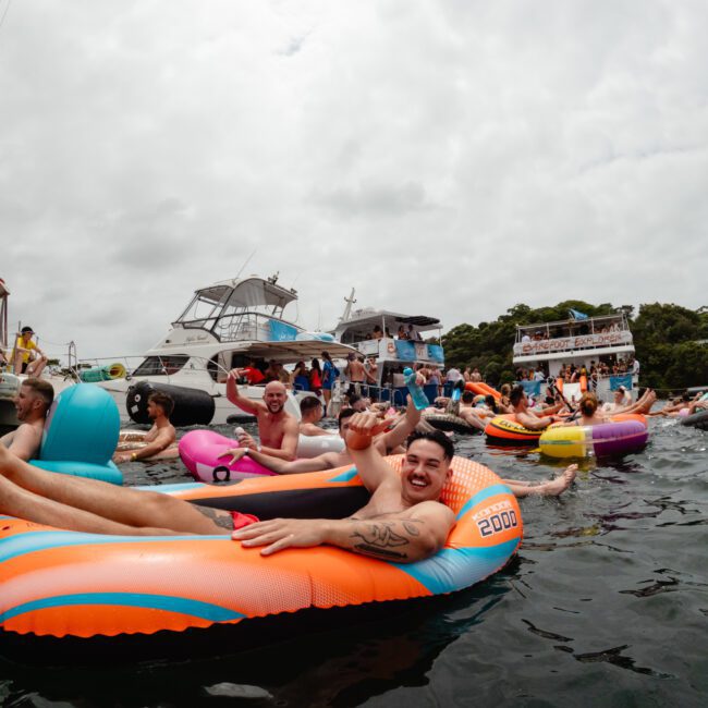 A group of people enjoying a sunny day on the water. They are floating on colorful inflatable tubes and rafts near several anchored boats from The Yacht Social Club. The atmosphere seems festive and relaxed, with one person prominently raising a beverage can and smiling at the camera.