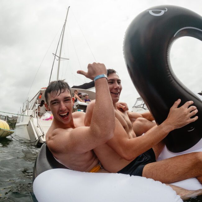 Two young men are sitting on a large inflatable swan in the water, smiling and making hand gestures. They appear to be enjoying themselves near a docked sailboat under a cloudy sky. Both wearing swim trunks and having a joyful time, they look ready for a Sydney Harbour Boat Hire Yacht Social Club event.