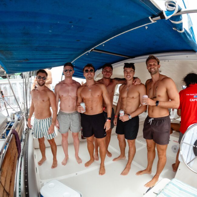 A group of six shirtless men in swim trunks stand on a boat's deck under a blue canopy, smiling and holding drinks. Another person in a red shirt labeled "Captain" stands at the steering wheel in the background. The scene appears to be sunny and festive, capturing the vibe of Boat Parties Sydney The Yacht Social Club.