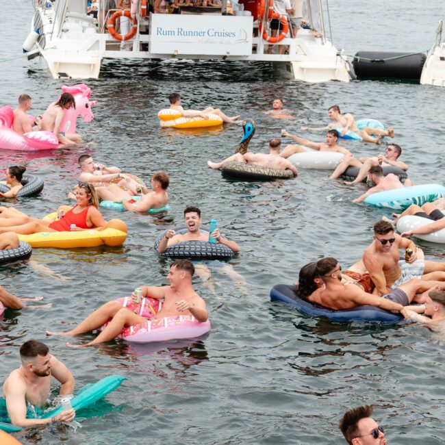 People are floating on inflatable rafts in the water near a boat labeled "Rum Runner Cruises." They are relaxing and enjoying the sunny day, with various colorful inflatables such as a pink flamingo and a yellow doughnut. Trees line the background, creating an idyllic setting for The Yacht Social Club's Sydney Harbour Boat Hire.