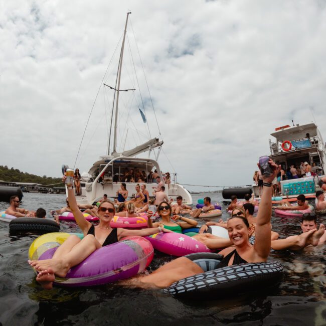 A group of people relaxing on colorful inflatable tubes in the water near anchored sailboats. They appear to be enjoying a sunny day, with some raising their drinks in celebration, while others chat and smile. The Yacht Social Club Sydney Boat Hire adds to the festive atmosphere with additional guests on board.