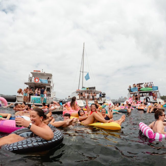 A group of people relaxing on various colorful inflatable rafts and floaties in the water near boats. The scene is festive with many smiling individuals, drinks in hand, and a backdrop of several anchored boats and a partly cloudy sky. Experience this joy at The Yacht Social Club Sydney Boat Hire.
