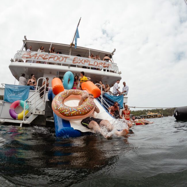 Adults and children slide down an inflatable slide from a two-story boat named "Barefoot Explorer" into the water. The boat, perfect for parties hosted by The Yacht Social Club Sydney Boat Hire, is decorated with colorful inflatables. Several people watch and enjoy the activity while a sailboat floats nearby.