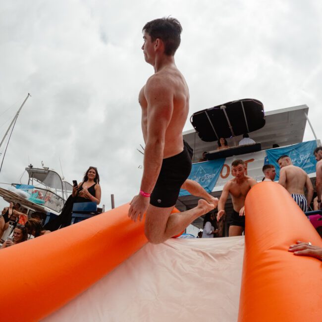 A man in black swim trunks slides down an inflatable orange and white slide on a boat from The Yacht Social Club. Others watch the action while someone photographs the scene. The sky is overcast, and the boat is docked near several others, perfect for unforgettable boat parties in Sydney Harbour.