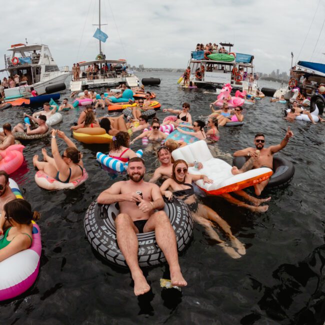 A lively group of people enjoying a party on a lake with several colorful floats and inflatable animals. Many are swimming, lounging, and interacting in the water, while a few others are on the boats in the background. The atmosphere is festive and cheerful, reminiscent of The Yacht Social Club Sydney Boat Hire events.