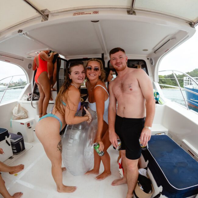 A group of people in swimsuits are gathered on a boat. Three individuals at the center smile at the camera; two women hold drinks, one man holds a bottle. Various items, including a life ring and coolers, are visible on deck. It's a sunny day by the water—a perfect setting for The Yacht Social Club event in Sydney Harbour.