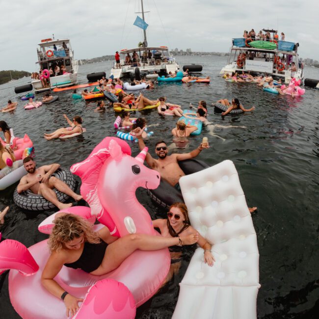 A lively scene of people enjoying a lake party. Many are in the water on colorful inflatable floats, like a pink unicorn and an ice cream cone. Luxury yachts from The Yacht Social Club are anchored nearby, with more people onboard. The sky is partly cloudy.