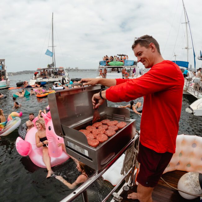 A man in a red shirt stands on a boat's deck, grilling burgers while various people on inflatable floats enjoy the water around the boat. The scene is lively with multiple boats and people in the background, epitomizing a typical day with The Yacht Social Club Sydney Boat Hire.