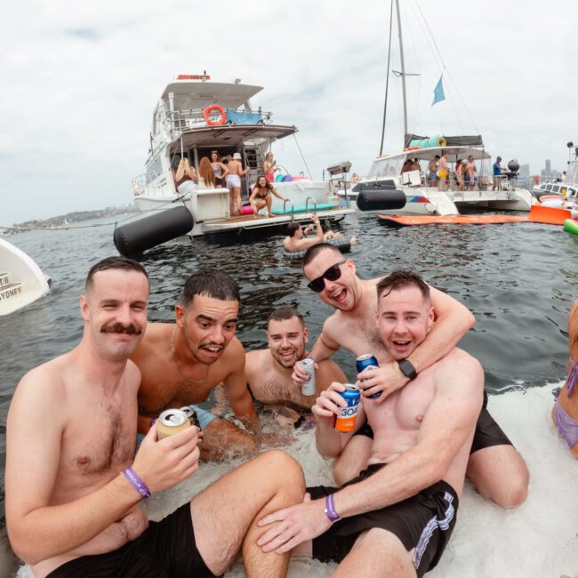 A group of five men, some holding drinks, are smiling and posing for a photo while sitting in the water near The Yacht Social Club Sydney Boat Hire. Several boats and people are visible in the background, enjoying a lively atmosphere under a cloudy sky.