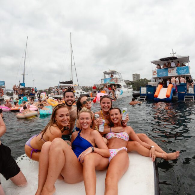 Several people are enjoying a day on the water, with a group of five individuals posing and smiling at the camera. They are sitting on the edge of a boat from Sydney Harbour Boat Hire The Yacht Social Club with drinks in hand, while additional people and boats are visible in the background with floaties and slides.