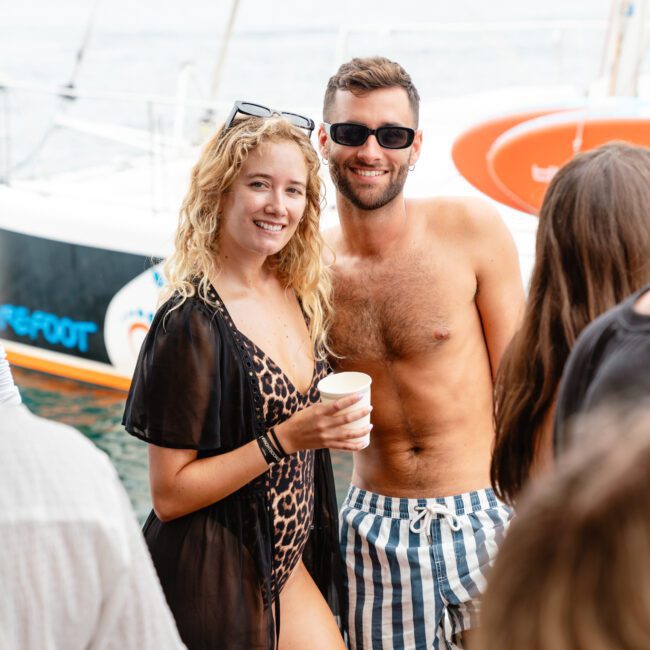 A man and woman in swimwear smile at the camera on a boat. The woman, in a leopard print swimsuit and black cover-up, holds a cup. The man, wearing striped swim trunks and sunglasses, has his arm around her shoulder. Boat parties by The Yacht Social Club Sydney can be seen in the background with other people having fun.