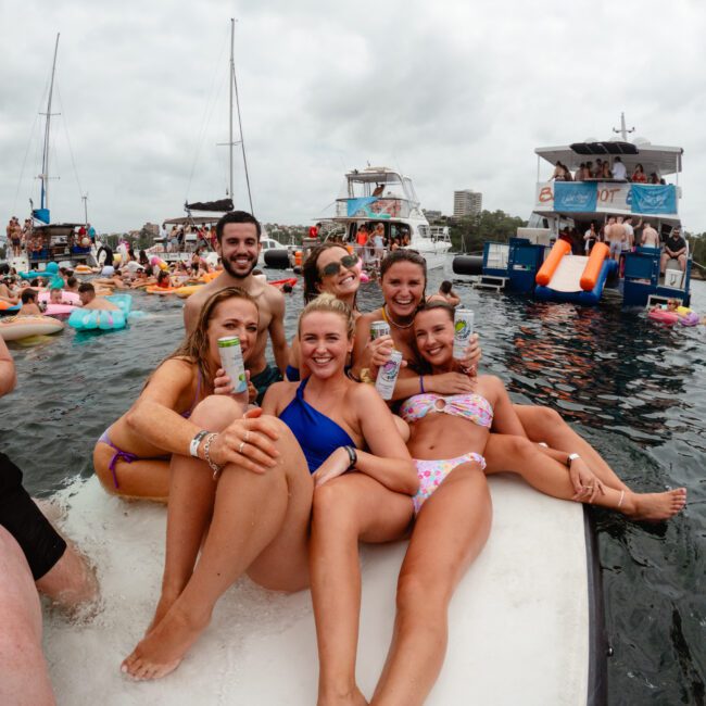 A group of six smiling people relaxes on an inflatable raft in the water, holding drinks and posing for a photo during The Yacht Social Club Event. Many other people and inflatable rafts are visible in the background along with boats and a cloudy sky. The atmosphere appears festive and lively.