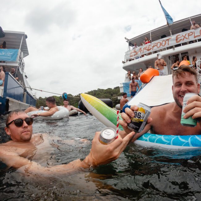 Two men in swimsuits float in the water near a boat labeled "Barefoot Explorer." They hold up cans of drinks and smile at the camera. Behind them, people are swimming and enjoying the scene, with some on inflatable pool floats and others on the boat. Experience fun with Sydney Harbour Boat Hire The Yacht Social Club!