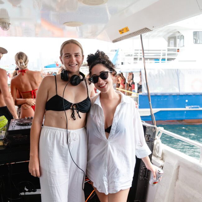 Two individuals are on a boat, posing and smiling at the camera. One is wearing headphones, a black bikini top, and white pants, while the other sports a white shirt and sunglasses. The lively scene showcases Boat Parties Sydney The Yacht Social Club with other people and boats in the background.