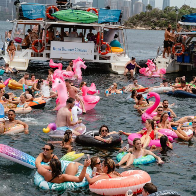A lively scene of people enjoying a sunny day in the water, surrounded by an array of colorful inflatable floats like flamingos and unicorns. Two boats from The Yacht Social Club Sydney Boat Hire, filled with more partygoers, are docked nearby, with the cityscape visible in the background.
