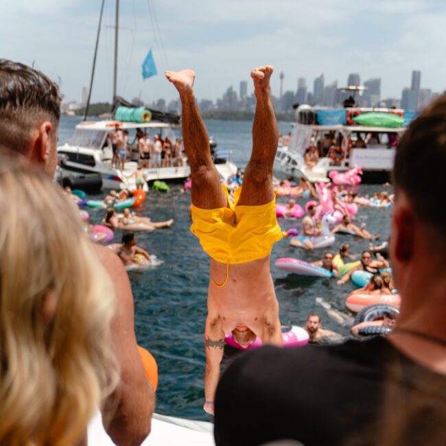 A person in yellow swim trunks performs a handstand on the edge of a boat. The surrounding boats and people are part of the vibrant Sydney Harbour Boat Hire The Yacht Social Club scene. Colorful inflatables float nearby, with the city skyline visible under a partly cloudy sky.