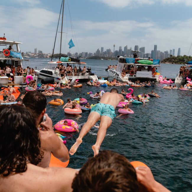 A lively scene of several boats gathered on a sunny day, with numerous people swimming and floating on colorful inflatables in the water. The backdrop includes a city skyline and a bridge, indicating an urban coastal area, perfect for Luxury Yacht Rentals Sydney.