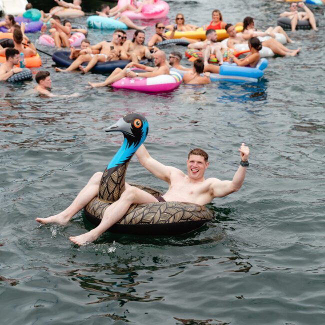 A man smiling and giving a thumbs-up while sitting on a bird-shaped pool float in a lake, surrounded by other people relaxing on various inflatables. The atmosphere is festive and fun, reminiscent of the vibrant gatherings hosted by Sydney Harbour Boat Hire The Yacht Social Club.