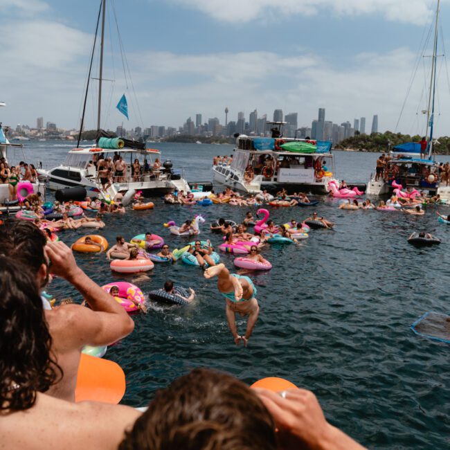 A lively scene of people enjoying a party on the water with The Yacht Social Club. Multiple boats are anchored close together, with many individuals swimming or relaxing on inflatable toys of various shapes and colors. A city skyline and the Sydney Opera House are visible in the background.