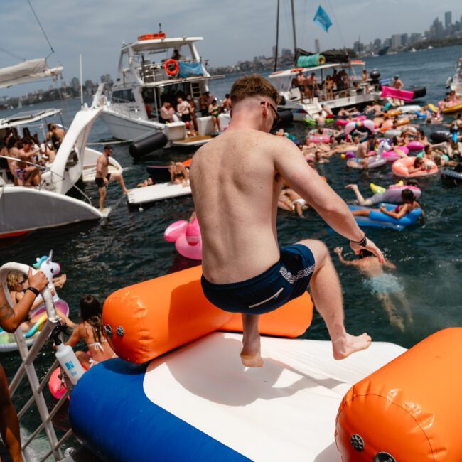 A shirtless man in blue swim trunks jumps off an inflatable platform into the water. Around him, numerous people are swimming and floating on colorful inflatable pool toys. Several boats from Sydney Harbour Boat Hire The Yacht Social Club are anchored nearby, with a city skyline visible in the background.