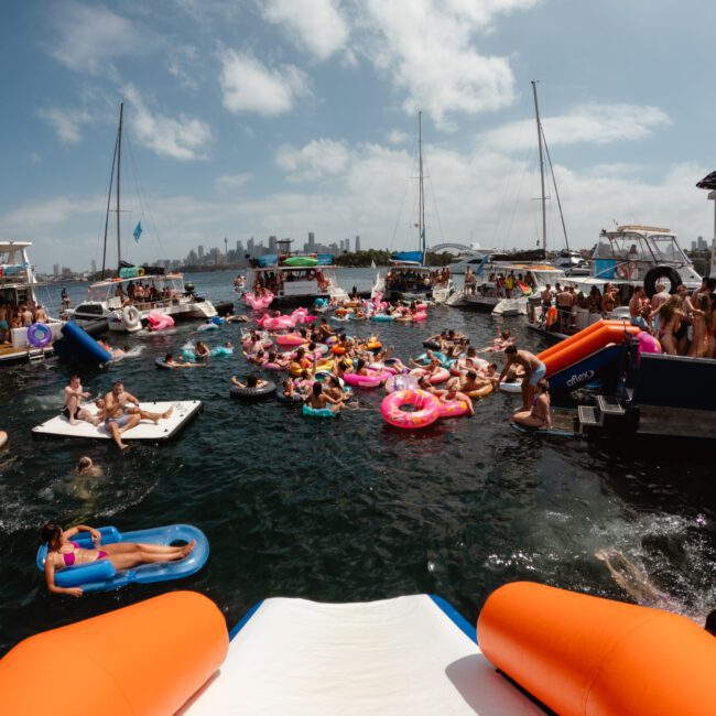 A lively scene of people enjoying a sunny day on the water. Various boats, including those from The Yacht Social Club Sydney Boat Hire, are anchored close together, with many individuals floating on colorful inflatables and rafts. The sky is partly cloudy, and a city skyline can be seen in the background.