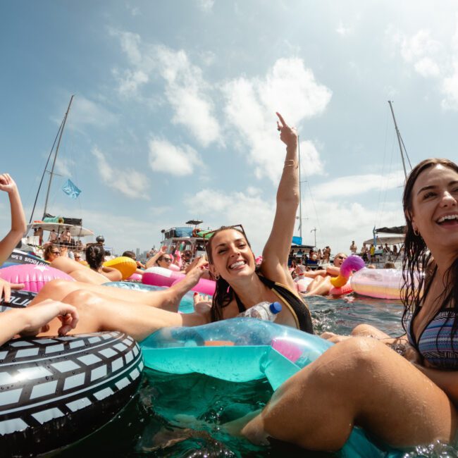 A group of young women enjoy a sunny day at a pool party, lounging on colorful inflatable tubes. They are all smiling, and one woman is raising her arm in excitement. In the background, more people relax on pool floats and boats. The Yacht Social Club Sydney Boat Hire adds to the lively atmosphere under the partly cloudy sky.