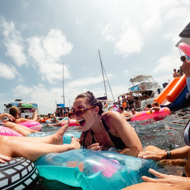 A person laughs energetically while lounging on an inflatable raft in a busy water area filled with others on flotation devices. Boats are visible in the background under a sunny sky, capturing the festive spirit of The Yacht Social Club Event Boat Charters.
