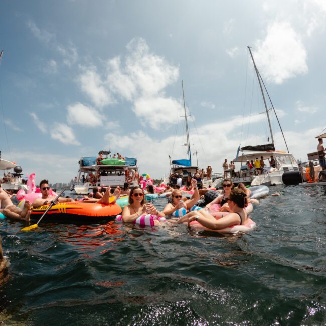 A group of people relax on inflatable floats in the water, surrounded by several anchored boats on a sunny day. They appear to be enjoying a festive gathering, some waving and others lying back under the clear blue sky, showcasing the epitome of Sydney Harbour Boat Parties with The Yacht Social Club.