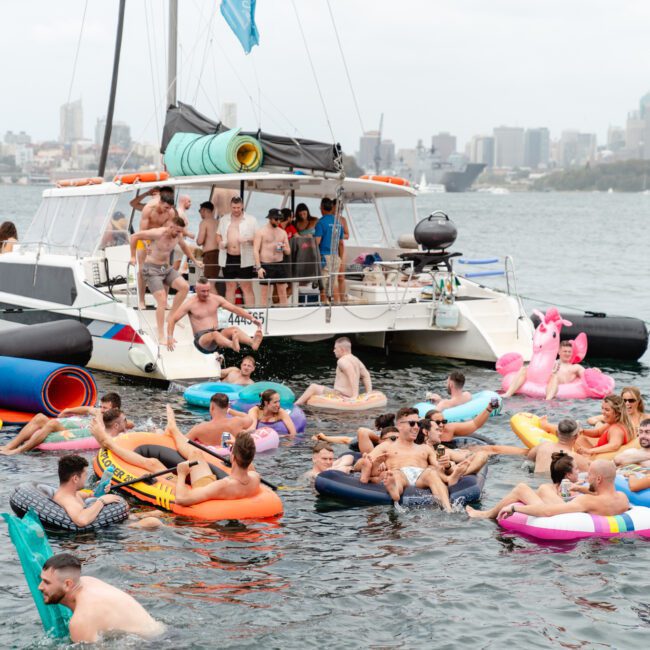 A group of people enjoy a sunny day on the water, lounging on inflatable floats in various shapes and colors. A large boat from The Yacht Social Club Sydney Boat Hire is anchored nearby with more people relaxing on its deck. The backdrop features a city skyline with tall buildings.