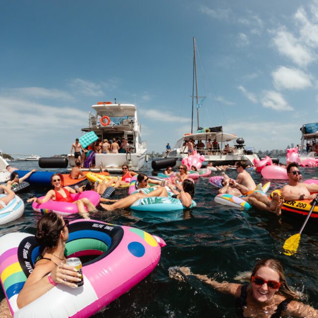 A lively scene of people floating on inflatable tubes and loungers in the water, surrounded by boats and yachts. The Yacht Social Club Event Boat Charters creates a relaxed and festive atmosphere as individuals enjoy the sunny day under the clear blue sky.