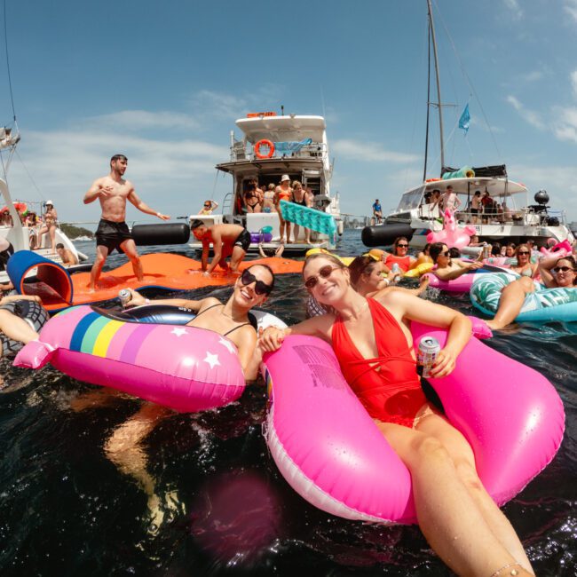 A lively group enjoys a day on the water, floating on colorful inflatable rafts near several boats. They're relaxing, laughing, and socializing under a bright, clear sky. Some hold drinks while others stand or swim in the water—an ideal scene from The Yacht Social Club's luxury yacht rentals in Sydney.